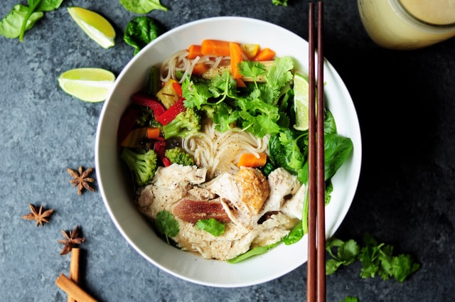 A bowl of rice noodles, chicken, veggies, and herbs.