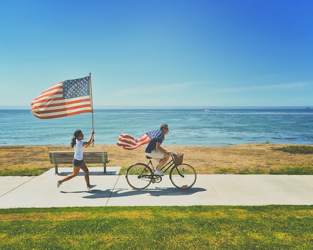 Man draped with a flag on a bike with a girl carrying a flag by the seaside.