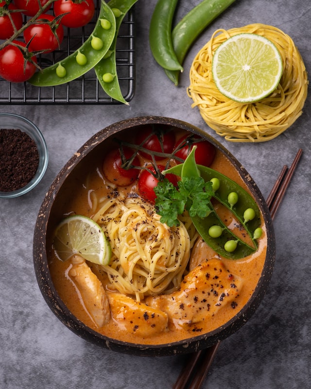 Bowl of chicken curry, noodles, and veggies on gray backdrop.