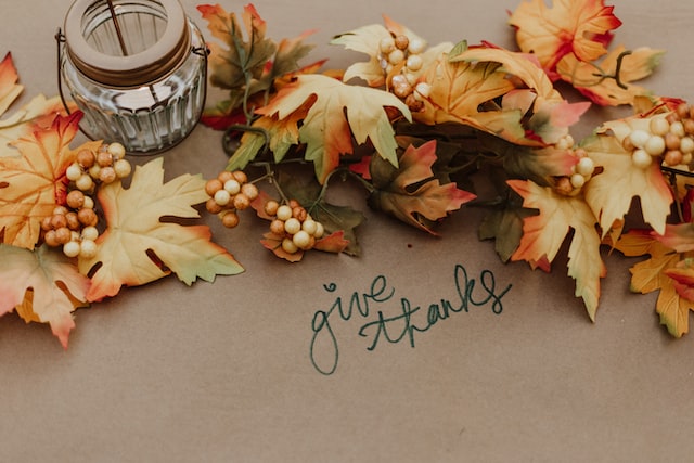 A cozy autumn scene with colorful leaves, a flickering candle, and the words "give thanks" on a table.