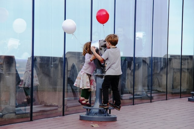Two children on a ledge, holding colorful balloons, with a telescope.